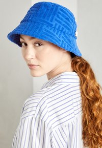 Chapeau seau bleu en tissu texturé avec un motif. Présente un bord festonné et une petite étiquette logo sur le côté.