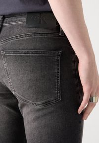 Černé džíny z denimu s přiléhavým střihem, s zadní kapsou a texturovanou koženou značkou Calvin Klein. Kovový prsten na ruce.