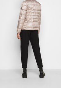 Světlý béžový péřový kabát s vodorovným prošívaným vzorem, který má strukturální boční panel a černé kalhoty. Černé kotníkové boty doplňují celkový vzhled.