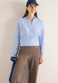 Femme portant un chemisier bleu clair à manches longues, un pantalon marron et une casquette bleue, tenant une veste en denim pliée, debout contre un mur beige.