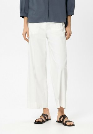 Person, die eine weiße Hose mit weitem Bein und schwarze flache Sandalen mit gekreuzten Riemen trägt und vor einem schlichten weißen Hintergrund steht.