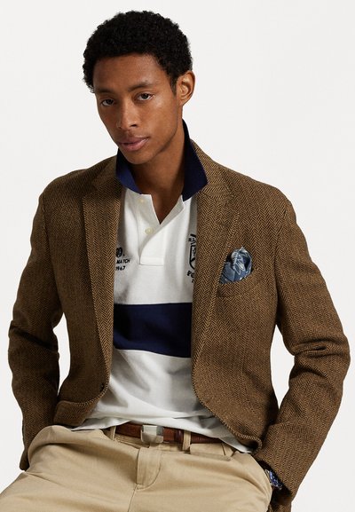 Bruine visgraat blazer over een wit en marineblauw gestreepte polo. De blazer heeft een notch-revers, terwijl het pochet blauw is met een bloemenpatroon. Beige broeken.