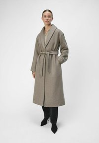 Abrigo largo de lana color marrón claro con cuello con muescas, cintura ceñida con cinturón y abertura frontal. Combinado con jeans oscuros y tacones negros de punta.