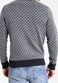 Pull en maille gris à motif avec un design en losange, poignets et ourlet côtelés, porté avec un jean bleu clair. Vue de dos montrant l'encolure et la texture.