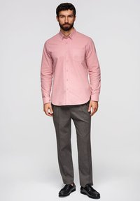 Homme barbu portant une chemise rose à boutons, un pantalon gris et des chaussures habillées noires, debout devant un arrière-plan clair uni.