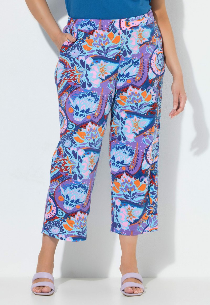 Mujer con pantalones anchos vibrantes con un patrón floral y abstracto colorido en púrpura, azul, naranja y rosa, combinados con sandalias lila.