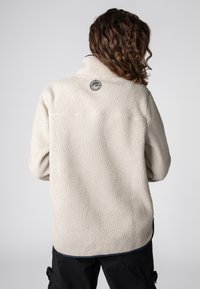 Cremfarbener Fleece-Pullover mit hohem Kragen, strukturierter Oberfläche und einem runden Logopatch auf der Rückseite, mit schwarzem Saum.