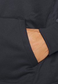 Sweat à capuche gris foncé en tissu doux et texturé. Il comporte une poche avant avec une main posée à l'intérieur, mettant en valeur l'ouverture de la poche et les détails de couture.