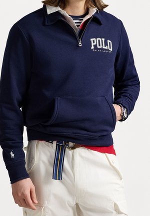 Personne portant un pull Polo Ralph Lauren bleu marine avec une fermeture éclair jusqu'au quart, un col blanc, un pantalon cargo blanc et une ceinture rayée, main dans la poche.
