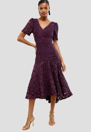 Mujer vistiendo un vestido midi de encaje morado con mangas cortas abullonadas y tacones altos dorados con tiras, de pie contra un fondo liso.