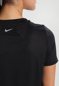 Camisa atlética negra con cuello redondo, mangas cortas y paneles de malla. Logo de Nike en plata prominente en la parte superior de la espalda. Tejido suave y ajustado.