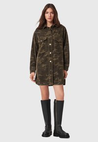 Frau, die eine lange, camouflagemusterte Cordjacke mit Vorderknöpfen und zwei Brusttaschen trägt, kombiniert mit schwarzen kniehohen Stiefeln mit Reißverschlüssen.