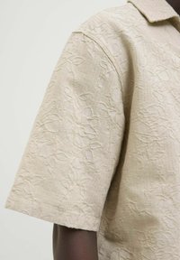 Camicia smanicata beige con un motivo floreale testurizzato, caratterizzata da un colletto e un fit rilassato.