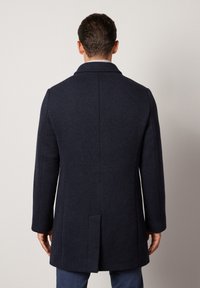 Marinblå ullrock med rak skärning, stor krage, långa ärmar och en slits bak. Minimalistisk design utan synliga knäppningar.