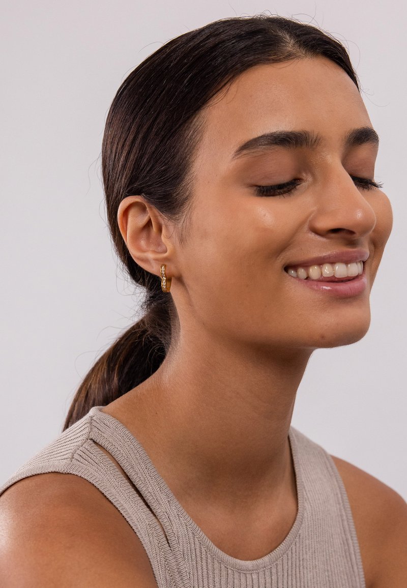Goldene Creolenohrringe mit strukturierter Oberfläche und funkelnden Akzentdetails, getragen von einer Person mit dunklen Haaren in einem neutralen Oberteil.