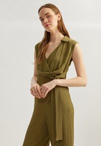 Olivgrön, ärmlös jumpsuit med omslag, krage, texturerad tyg och ett knyt i midjan. Bred benprofil.