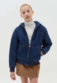 Sudadera con cremallera de mezclilla en azul oscuro, con capucha, bolsillos frontales, puños de canalé en azul marino y un corte ligeramente corto, combinada con una falda de pana beige.