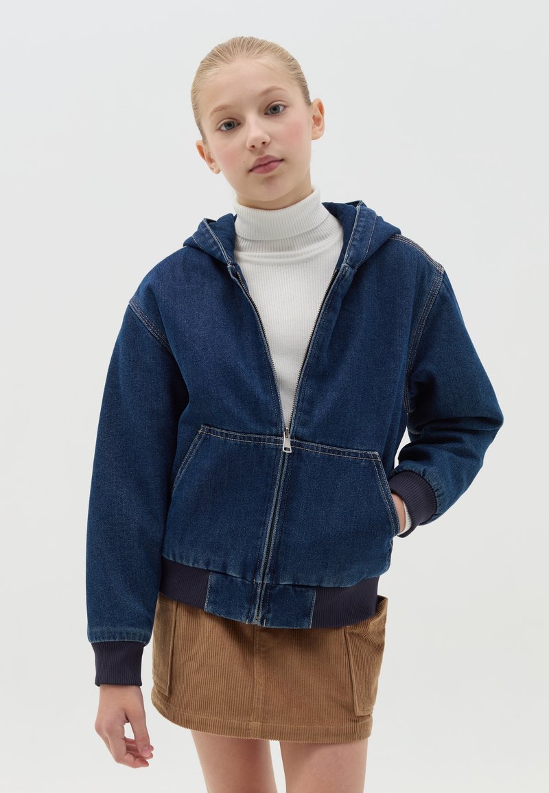 Sudadera con cremallera de mezclilla en azul oscuro, con capucha, bolsillos frontales, puños de canalé en azul marino y un corte ligeramente corto, combinada con una falda de pana beige.
