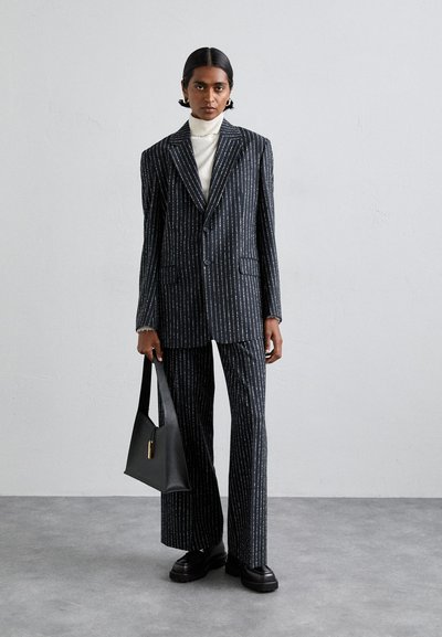 Σκούρο ριγωτό κοστούμι με στενές λευκές γραμμές, κομμένο για τέλεια εφαρμογή, σακάκι μονής κουμπώματος, ταιριαστά παντελόνια, συνδυασμένο με αναγεννησιακό πουλόβερ και μαύρη τσάντα.