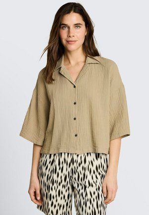 Kobieta z długimi brązowymi włosami, ubrana w beżową bluzkę zapinaną na guziki oraz czarno-białe spodnie w cętki, stojąca na jednolitym tle.