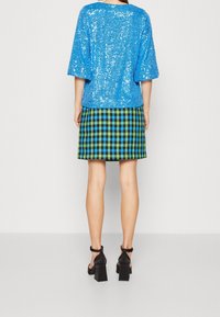 Top blu con paillettes e maniche oversize abbinato a una gonna a quadri houndstooth nera e verde. Sandali neri con tacco a blocco completano l'outfit.