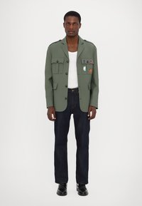 Veste en tissu vert avec quatre poches avant, des patches décoratifs et des boutons, portée sur une chemise blanche et un jean en denim foncé. Chaussures noires.