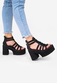 Sandalias de plataforma negras con múltiples correas, cierre con cremallera en la parte posterior y tacones anchos. Combinadas con unos jeans desgastados de un azul claro.