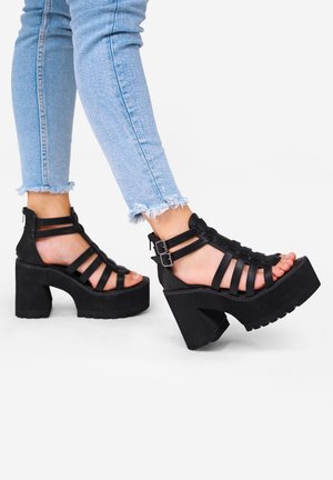 Sandalias de plataforma negras con múltiples correas, cierre con cremallera en la parte posterior y tacones anchos. Combinadas con unos jeans desgastados de un azul claro.