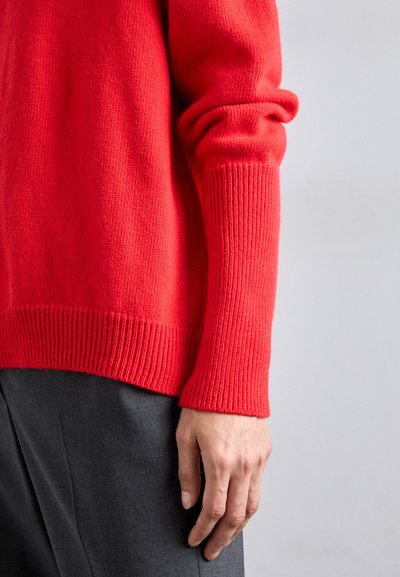 Bright red sweater with ribbed cuffs and hem, made from soft knit material, worn over dark gray trousers, showing a close-up of the sleeve.