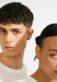 Close-up van een man en vrouw met kort donker haar, die witte tops dragen en bijpassende kleine ronde oranje oorbellen, tegen een effen achtergrond.