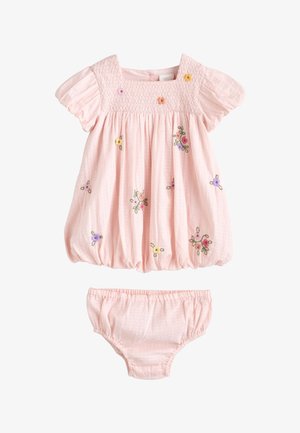 Barn iført en lyserød kjole med puffede ærmer, med en smocket krop og farverige broderede blomster på bryst og skørt.