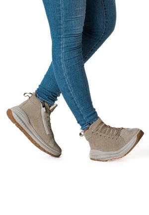 Person wearing light brown high-top sneakers with thick soles and blue skinny jeans, standing with one foot slightly raised.