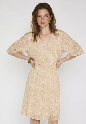 Donna con capelli castani ondulati che indossa un abito floreale rosa chiaro al ginocchio, con maniche corte, orecchini a cerchio dorati e braccialetti, in piedi con le mani sui fianchi.