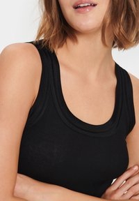 Tuva sieviete ar īsiem brūniem matiem, valkājot melnu bezpiedurkņu ribu topu, rokās saliktām krūtīs un lūpām nedaudz atvērtām.