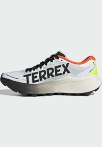 Chaussure de course avec une tige blanche, des lacets noirs et un grand logo "TERREX". Présente un accent orange au niveau du talon et une semelle noire épaisse.