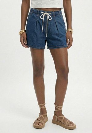 Naine, kes kannab siniseid teksapükse valge nööriga, beeže rihmikuga sandaale, valget pitsilist pluusi, kuldseid käevõrusid ja kuldset käekella.