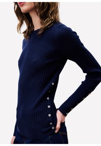 Frau, die einen marineblauen gerippten Pullover mit Perlenknöpfen entlang der Seitennaht und dem Ärmel trägt, kombiniert mit dunklen Jeans.