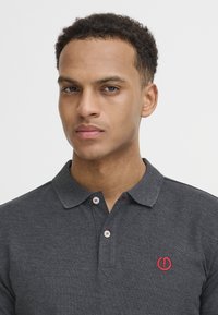 Kulgrå polo-shirt med krave, der har røde detaljer på knapperne og et rødt logo på brystet. Blød tekstur.