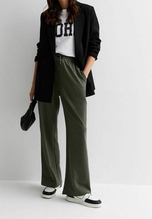 Kobieta ubrana w czarną marynarkę, białą koszulkę z nadrukiem "OH", zielone spodnie o szerokich nogawkach, czarno-białe trampki, trzyma czarną teksturowaną torbę.