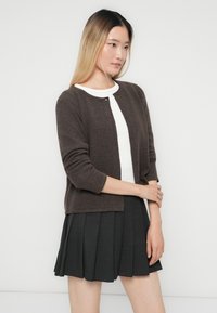 Donna che indossa un cardigan marrone scuro sopra una maglietta bianca e una gonna plissettata nera, in piedi con le braccia incrociate contro uno sfondo chiaro e semplice.