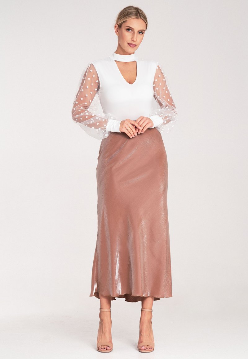 Weißes Oberteil mit einem Ausschnitt mit Schlüsselöffnung und transparenten, gepunkteten Ärmeln. Brauner, glänzender Midirock mit leichtem Fall. Einfache, nudefarbene Absätze.