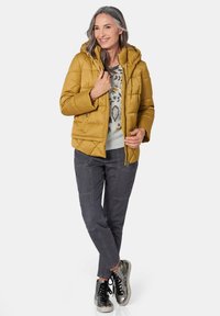 Gelbe Daunenjacke mit Kapuze, Reißverschluss und seitlichen Taschen. Getragen über einem gemusterten grauen Pullover und kombiniert mit grauen Hosen und metallischen Sneakers.