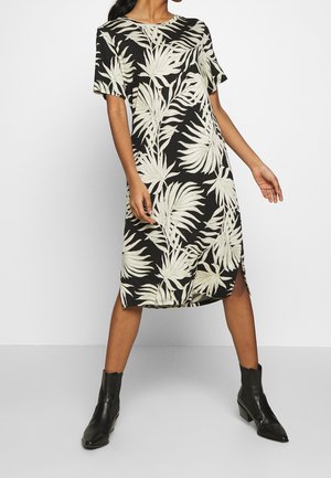 Woman wearing a black knee-length dress with large white tropical leaf patterns and black ankle boots, posing with legs apart.