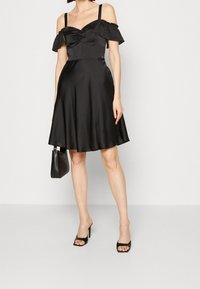Černé saténové šaty mají přiléhavý živůtek s volánkovými rukávy odhalenými na ramenou, rozšířenou sukni a hladkou texturu. Doplněné o černou kabelku.