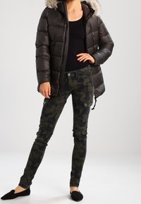 Černá zateplená bunda s umělou kožešinou na kapuci, spárovaná s maskáčovými cargo kalhotami a černými plochými botami. Hladká textura, přiléhavý střih.