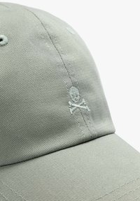 Lys grå kasket med et struktureret stof. Har små broderier af døskalle og korslagte knogler i matchende tråd foran. Bøjet skygge design.
