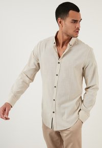 Camisa beige de botones, fabricada en un tejido ligero, con cuello extendido, mangas largas y botones oscuros, con un dobladillo curvado y una textura sutil.