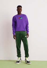 Lilla sweatshirts med Nike-logo, parret med grønne velour-joggingbukser med sidelister og sorte sneakers med neonlys.