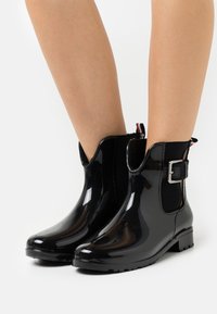 Bottes de cheville en caoutchouc noir avec une finition brillante, des panneaux latéraux élastiques et un détail décoratif en boucle argentée. Semelles texturées pour une meilleure adhérence.