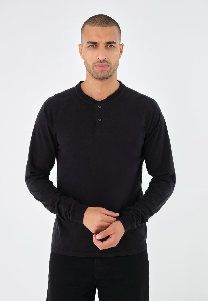 Schwarzes Langarm-Henley-Shirt mit Rundhalsausschnitt, ausgestattet mit einer Dreiknopfleiste und gerippten Bündchen, getragen mit dunklen Hosen.